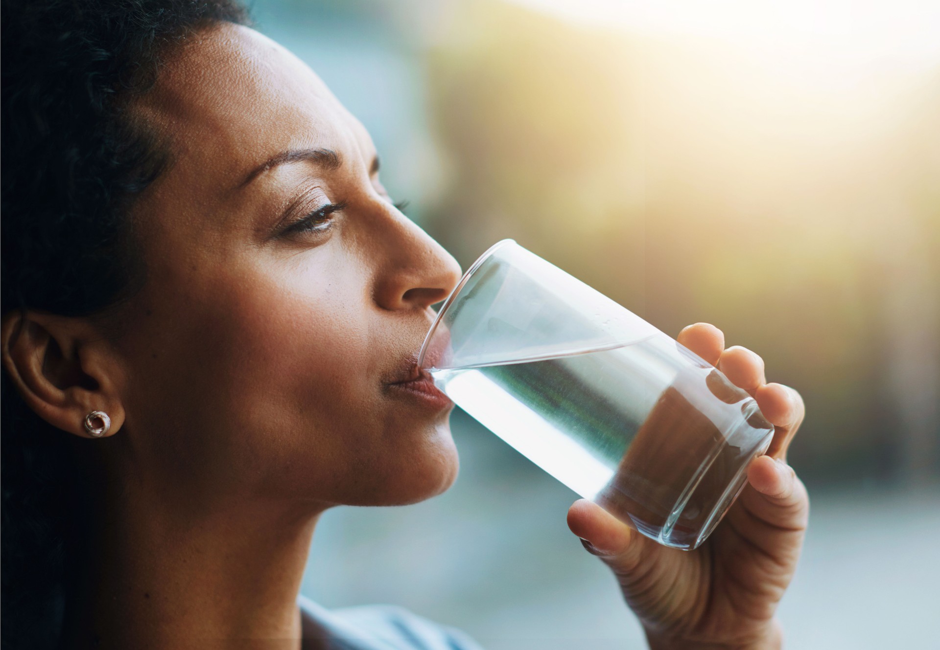 Woman, thinking and home with drinking water in glass for wellness, wellbeing and hydration. Profile, female person and satisfied with self care for thirst, health and clam energy in lens flare